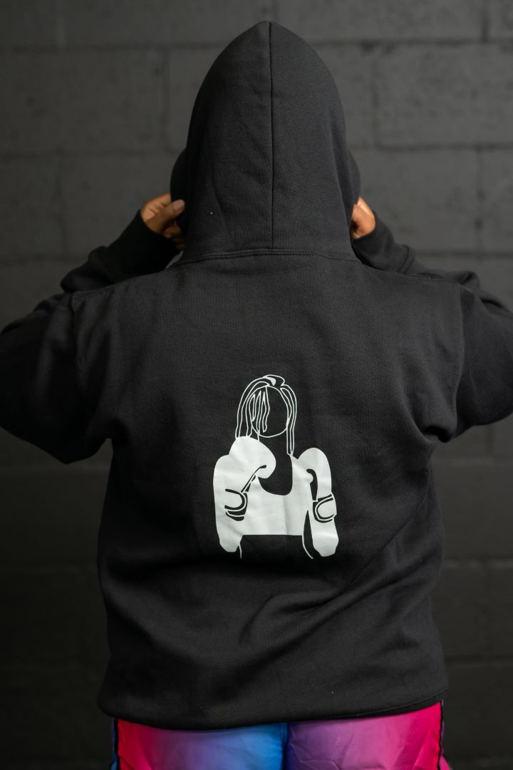 Model has her back turned towards the camera with the hood of the hoody pulled up. The hoody is all black with an off white glow in the dark female fighter silhouette centered on the back. The silhouette is a faceless woman with boxing gloves and a sports bra on with her hands up in a fighting stance.