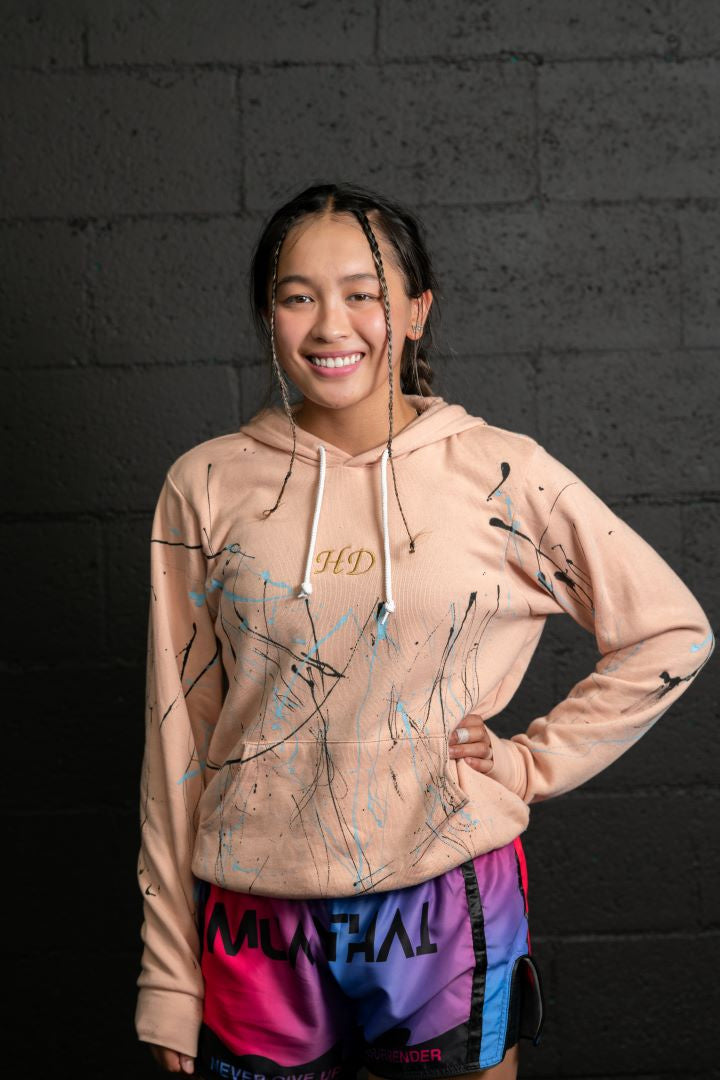 Model is looking at the camera smiling. She is wearing a pink hoody with black and light blue paint splatter. The letters HD are embroidered in gold and centered on the chest.