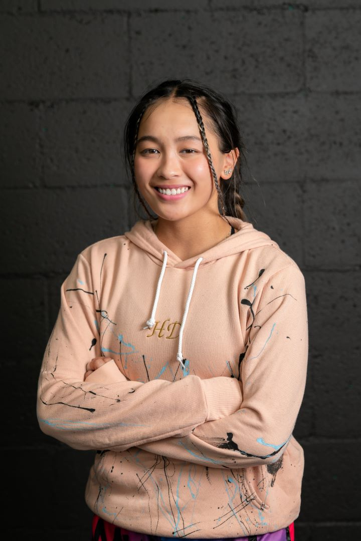 Model is looking at the camera smiling with her arms crossed. She is wearing a pink hoody with black and light blue paint splatter. The letters HD are embroidered in gold and centered on the chest.