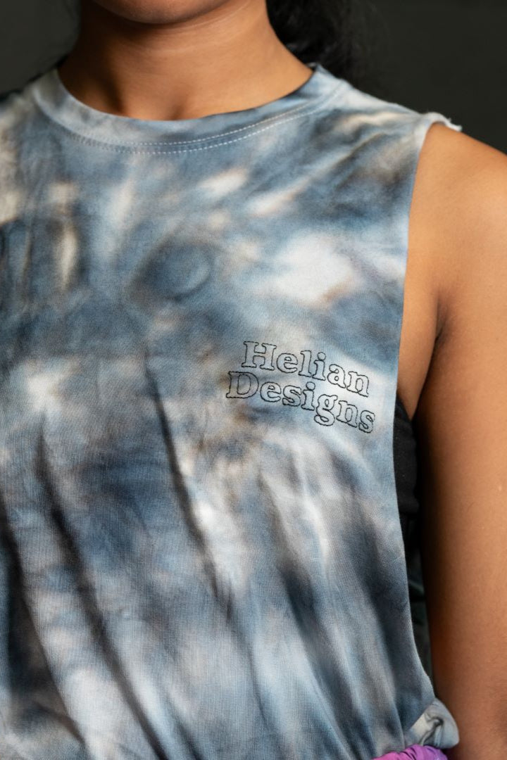 a close up shot of the tie die muscle t shirt. It is black spiral tie dye with the words Helian Designs stacked and centered on the left breast