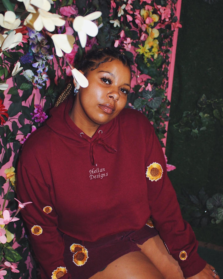 Female model in flower child cropped hoody. The hoody is burgundy, and the sleeves are embroidered with sunflowers. The company name Helian Designs is centered in white on the chest of the hoody.